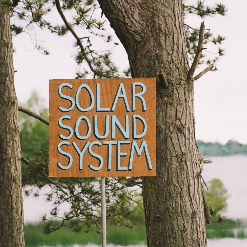 Wooden sign with the words 'Solar Sound System' in front of large trees and a lake